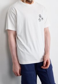 Man med en vit t-shirt som har tre små svart-vita cirkulära lappar på bröstet och marinblå byxor med fickor.