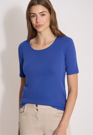 Femme aux longs cheveux bruns portant une chemise bleue ajustée à manches courtes et un pantalon beige clair, debout devant un fond clair uni.