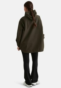 Femme aux cheveux tressés portant une veste à capuche oversize couleur olive, un pantalon noir évasé et des baskets blanches, debout, vue de dos.