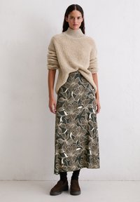 Un pull à col roulé beige et texturé associé à une longue jupe verte avec un motif floral blanc. Le modèle porte des bottes chelsea marron foncé.