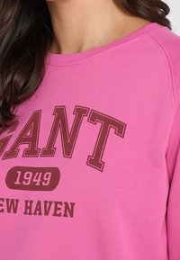 Sweat-shirt rose avec un col rond, des manches longues, et un motif à l'avant avec le texte "GANT 1949 NEW HAVEN" en marron foncé.