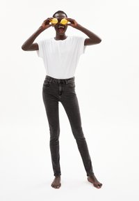 Schwarze hochgeschnittene Jeans und ein lockeres weißes T-Shirt. Die Person hält Zitronen vor ihr Gesicht und steht barfuß vor einem weißen Hintergrund.
