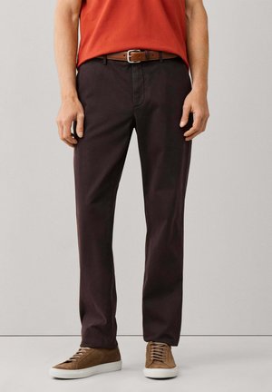 Hombre que lleva pantalones marrón oscuro, cinturón de cuero marrón, camisa naranja y zapatillas de ante marrón con suelas blancas, de pie contra un fondo liso.