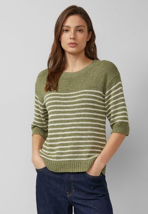 Kobieta o długich brązowych włosach, ubrana w oliwkowo-zielony i biały sweter w paski z dzianiny z rękawami do łokcia oraz ciemnoniebieskie dżinsy.