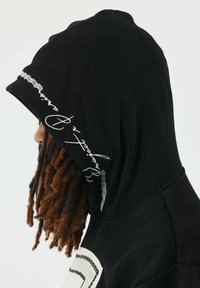Sudadera con capucha negra que presenta un diseño bordado texturizado a lo largo del borde de la capucha. Cuenta con una impresión gráfica en la parte frontal y un ajuste relajado.
