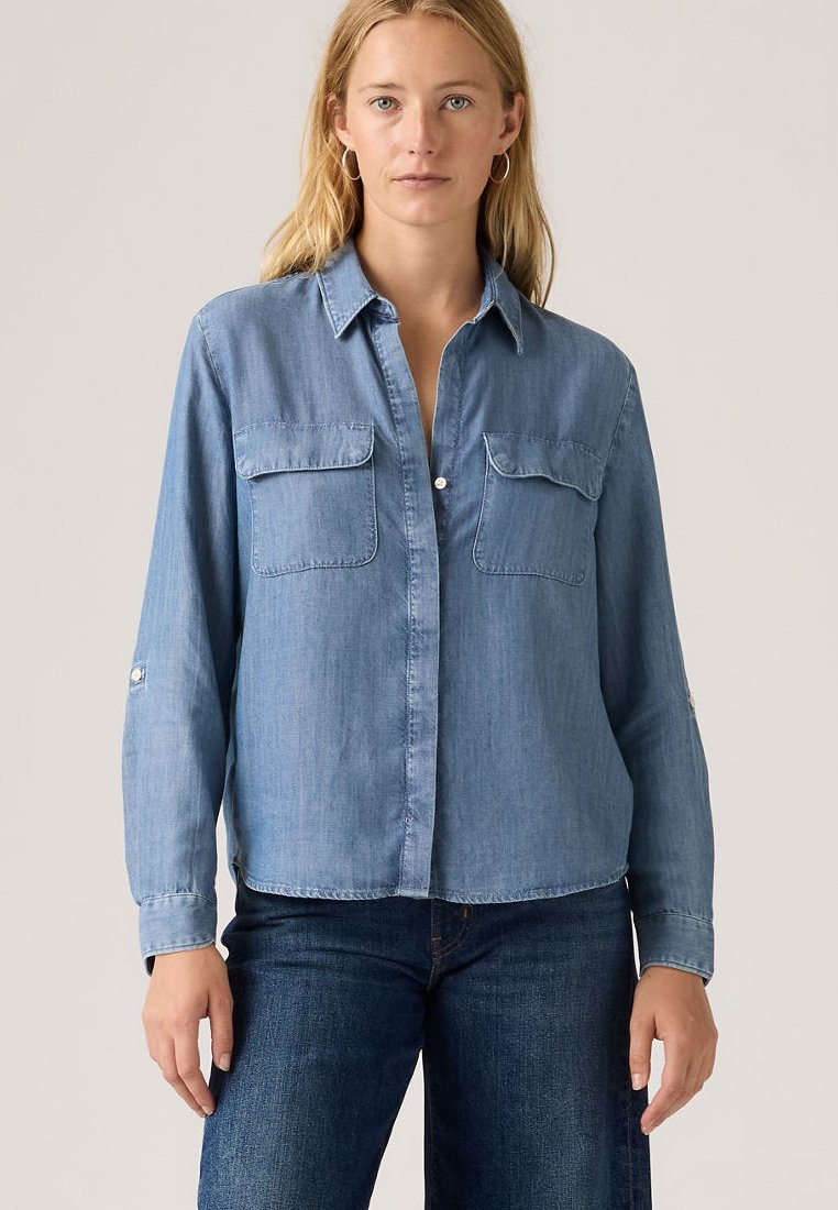 Kvinde iført lyseblå langærmet denimskjorte med brystlommer og mørkeblå jeans, stående mod en ensfarvet lys baggrund.