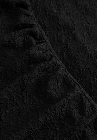 Čierna textúrovaná látka s jemnými vzormi, ktorá na jednej strane obsahuje zhromaždené volány. Povrch má mäkký, mierne vyvýšený pocit.