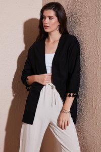 Cárdigan negro de delante abierto con mangas de tres cuartos y puños con borlas, combinado con pantalones de cintura alta en color beige claro. Fondo texturizado.