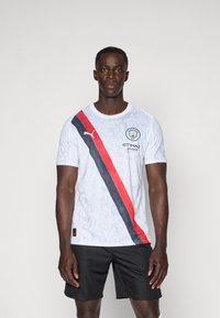 Bijela atletska majica s crvenom i crnom dijagonalnom prugom, s natpisom Manchester City i detaljima u sivoj boji. Uparen s crnim šortsevima.