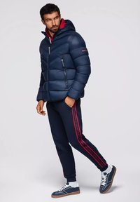 Giacca a vento blu navy con cappuccio e tasche con zip, abbinata a pantaloni jogger blu navy con strisce rosse ai lati e sneaker blu con dettagli bianchi.