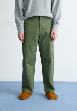 Person iført olivengrønne bukser med lige ben, grå langærmet top og brune ruskindssko med smutte-på-stil, stående på blåt gulv.