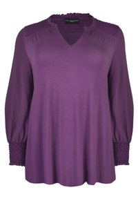 Lila pusero, jossa on V-pääntie, smockattu olkapäätyyli ja pitkät hihat. Valmistettu pehmeästä kankaasta, jossa on ker gathered-tiedot hihansuissa.