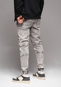 Persona que lleva puestos unos jeans de lavado ácido gris claro, zapatillas negras y blancas, calcetines blancos y una sudadera negra, de pie contra un fondo gris.