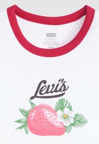 Camiseta blanca con cuello de canalé rojo, que presenta un gráfico de una fresa rosa y una flor blanca con hojas verdes, con el logo de la marca en la parte superior.