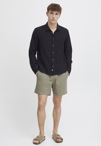 Sort skjorte med knapper og en lomme, kombineret med olivengrønne texturerede shorts. Modellen bærer brune sandaler og står med hænderne i lommerne.