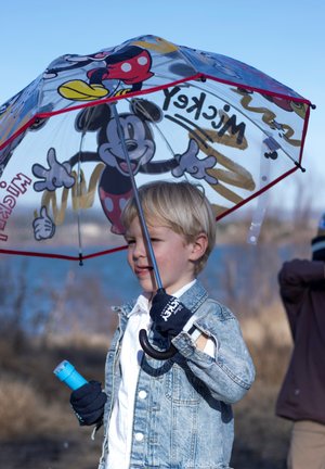 Ein Kind hält einen transparenten Regenschirm mit Mickey Mouse, trägt eine Jeansjacke, ein weißes Shirt und blaue Handschuhe und hält eine blaue Taschenlampe.
