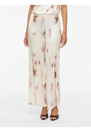 Kobieta w beżowych spodniach z szerokimi nogawkami i wysokim stanem, z abstrakcyjnymi brązowymi i fioletowymi kwiatowymi wzorami oraz czarnych sandałkach na obcasie z odkrytymi palcami.