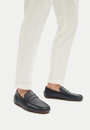 Mocasini din piele marine cu un aspect neted și cusături decorative, asortați cu pantaloni albi texturați, rulați la tiv.