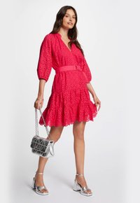 Vestido rosa brillante con detalles recortados, volantes en capas y cintura ceñida con cinturón. Combinado con sandalias de tacón cuadrado plateadas y un bolso acolchado.