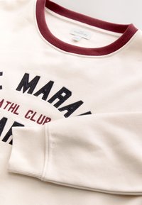 Sweat-shirt de maternité couleur crème avec un col bordeaux et un texte brodé en noir et bordeaux indiquant "Marathon Athl Club."