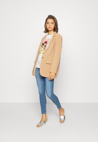 Blazer bege ajustado com um único botão dourado, combinado com uma t-shirt gráfica com arte colorida, jeans skinny de ganga e mules prateadas.