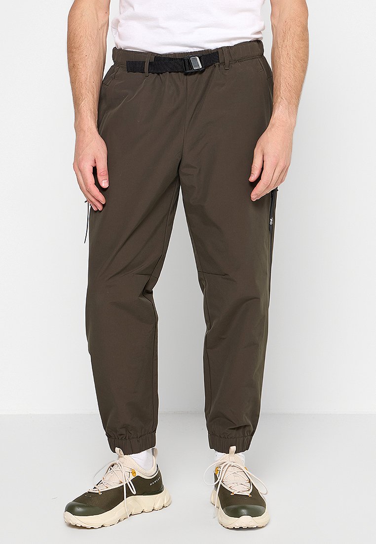 Uomo con pantaloni jogger marrone scuro con bordi elasticizzati, cintura nera, calze bianche e scarpe da ginnastica beige e verde oliva, in piedi contro uno sfondo bianco.