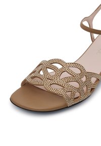 Sandalo beige con tomaia decorativa intagliata e dettagli di perline, suola piatta e cinturino alla caviglia regolabile. Texture liscia e design minimal.