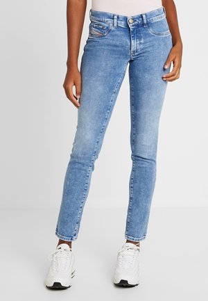 Person iført lyseblå skinny jeans og hvide sneakers, der står mod en ensfarvet lys baggrund.