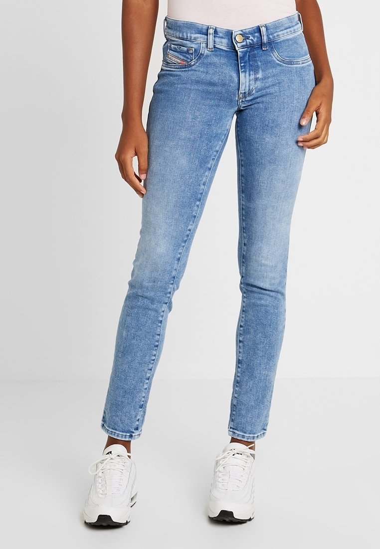 Personne portant un jean skinny bleu clair et des baskets blanches, debout devant un fond uni clair.