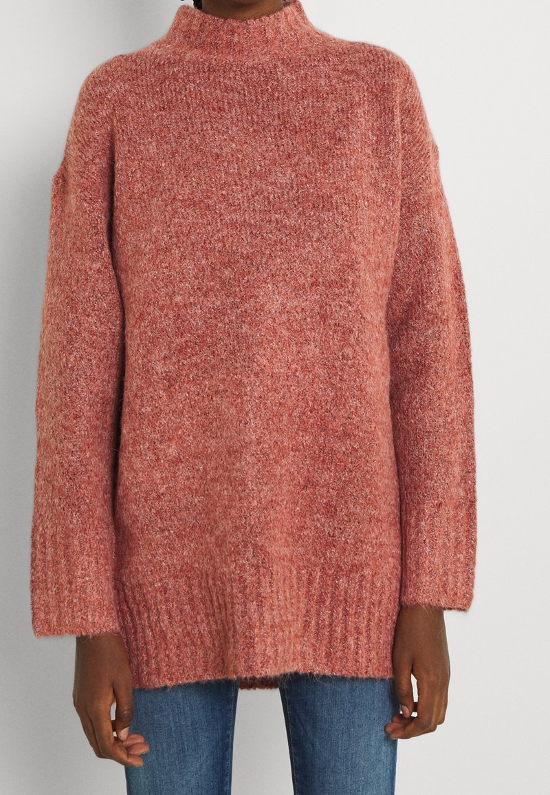 Persoon in een losse, textuurvolle, hooggesloten, stofroze trui en blauwe jeans, staande voor een effen witte achtergrond.