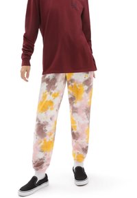 Tie-dye byxor i rosa, gult och brunt med avslappnad passform och elastiska mudd, tillsammans med en vinröd långärmad tröja och svarta slip-on skor.