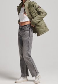 Grön militärjacka, vit kort topp, grå högmidjade jeans och vita sneakers med texturerade ytor och fransade kanter.