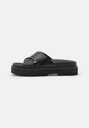 Sort platformsandal med brede teksturerede krydsede stropper og tyk rillede sål på ensfarvet baggrund.