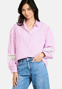Woman wearing loose pink and white striped button-up blouse with lace detail on sleeves and high-waisted blue jeans, standing with one hand in pocket.
