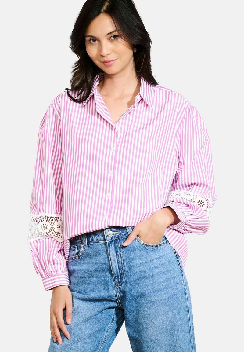 Woman wearing loose pink and white striped button-up blouse with lace detail on sleeves and high-waisted blue jeans, standing with one hand in pocket.