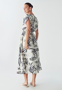 Cvečni midi obleka v krem barvi z modrimi botaničnimi motivi, kratkimi rokavi, elastičnim pasom in stranskimi razporki. Oblečena s ploščatimi sandali.