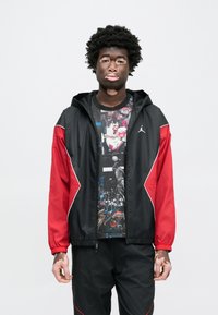 Αδιάβροχο μπουφάν μαύρο και κόκκινο με κουκούλα, με λευκές λεπτομέρειες. Κάτω από αυτό, ένα γραφικό T-shirt με εικόνες μπάσκετ. Φορεμένο με μαύρο παντελόνι.