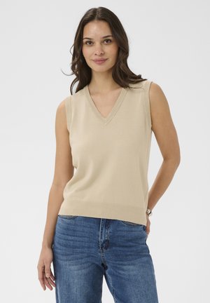 Giovane donna con capelli castani che indossa un maglione beige senza maniche a scollo a V e jeans blu, in piedi con una mano in tasca davanti a uno sfondo semplice.