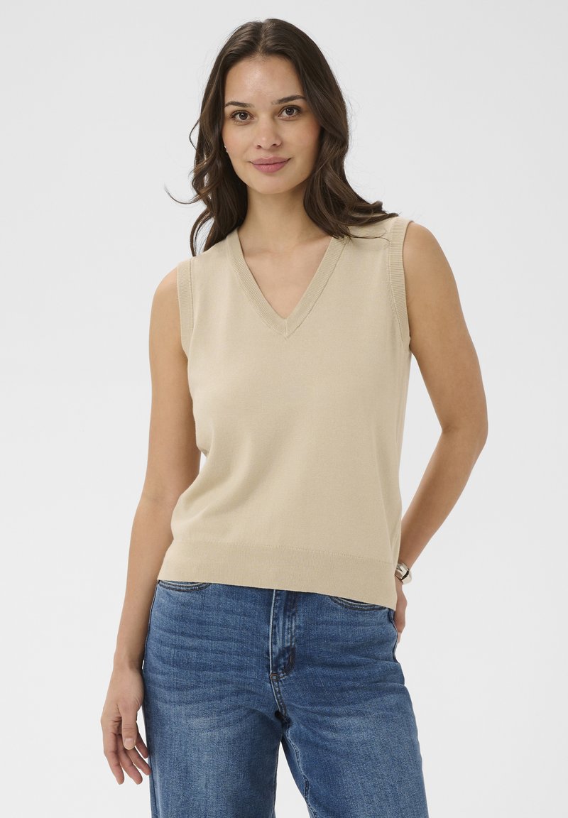 Jeune femme aux cheveux bruns portant un pull sans manches beige à col en V et un jean bleu, debout avec une main dans la poche devant un fond uni.