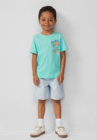 Turkusowa bawełniana koszulka z kolorowym nadrukiem przedstawiającym plażę i tekstem, w połączeniu z jasnoniebieskimi szortami dżinsowymi i białymi butami.