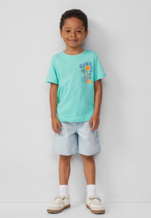 Türkises Baumwoll-T-Shirt mit einem bunten Druck eines Strandszene und Text, kombiniert mit hellen Jeansshorts und weißen Schuhen.