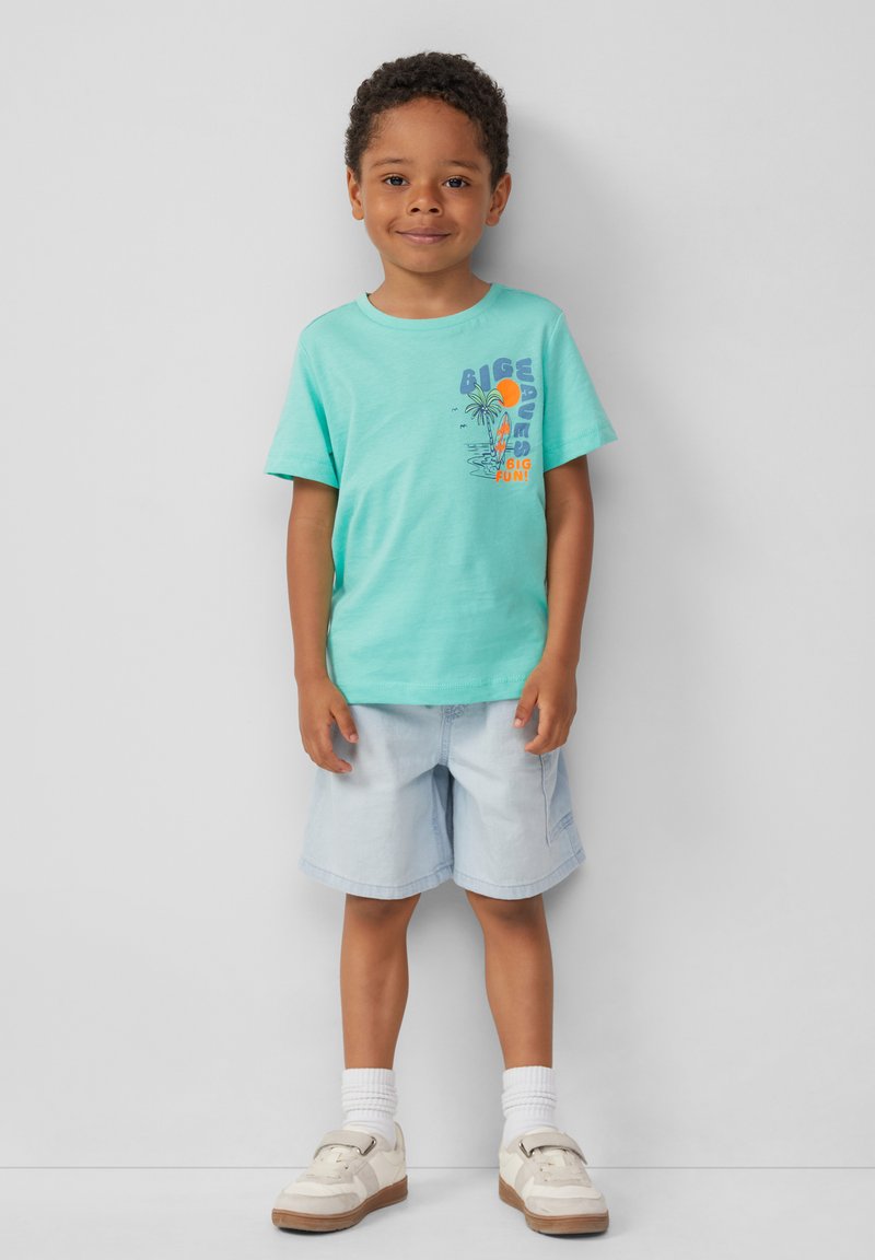Turkusowa bawełniana koszulka z kolorowym nadrukiem przedstawiającym plażę i tekstem, w połączeniu z jasnoniebieskimi szortami dżinsowymi i białymi butami.