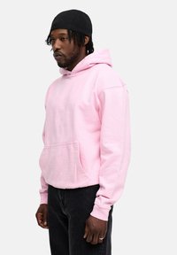 Pink hoodie with a front pocket and drawstring hood, made of soft fabric. Worn with black denim jeans. Black cap on head.
