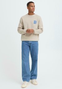 Beige genser med en blå brodert "B" på brystet, kombinert med vide, blå denimjeans og beige joggesko. Jevn tekstur.