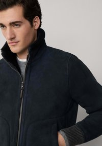 Mornarići suknja s visokim ovratnikom, zatvaranjem na patentni zatvarač i rebrastim sivim pletenicama; ima dvije prednje džepove i glatku teksturu.