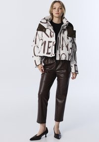 Jeune femme portant une veste courte en duvet blanche avec un texte abstrait marron, un pantalon en cuir marron foncé, un haut noir et des escarpins à talons kitten noirs.