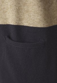 Pulôver de malha com um padrão de riscas em bege e azul marinho, apresentando um pequeno bolso na seção azul marinho, destacando o tecido texturizado.