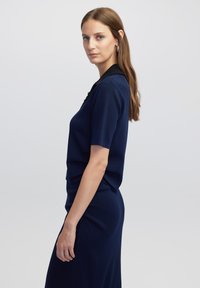 Žena s dlouhými hnědými vlasy, oblečená v námořnickém modrém triku s krátkým rukávem a odpovídající sukni, stojící bokem na světlém šedém pozadí.