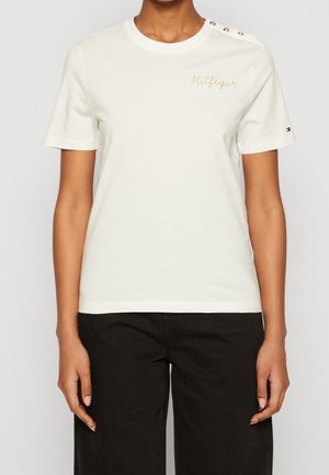 Persoon die een wit shirt met korte mouwen draagt met "Hilfiger" geborduurd in goud en een zwarte broek, staand tegen een effen achtergrond.