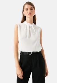 Weiße ärmellose Bluse mit plissee-vorderseitiger Gestaltung, hohem Kragen und verstecktem Knopfdetail, kombiniert mit schwarzen, hochgeschnittenen Hosen und einem Gürtel.